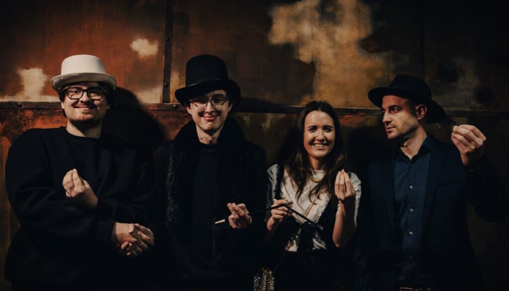 Four PredictLeads team members dressed in 1920s Gatsby-themed attire smiling and posing after completing a mystery escape game at Enigmarium. They wear vintage-style hats and accessories, standing against a rustic, dimly lit background.
