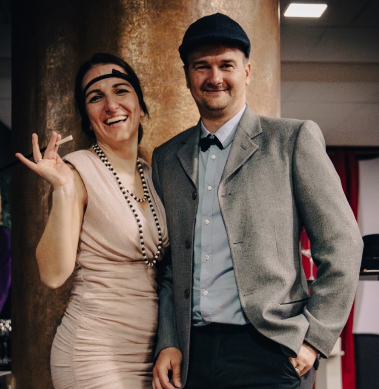 Smiling man and woman in vintage 1920s-style attire at a themed event, posing confidently in front of a gold column. The woman wears a flapper-style dress with pearls and a headband, while the man is dressed in a grey blazer, light blue shirt, and flat cap.