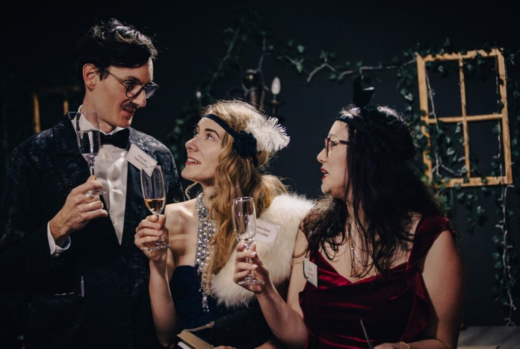 Three PredictLeads team members in elegant 1920s Gatsby-style costumes clinking champagne glasses during a themed mystery event. They are wearing vintage dresses, pearls, feathers, and headpieces, smiling and engaged in lively conversation.